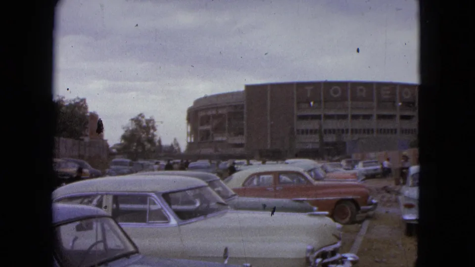 1962 Acapulco home-movie frame with parked sedans, pedestrians, and a large building carrying TOREO lettering.