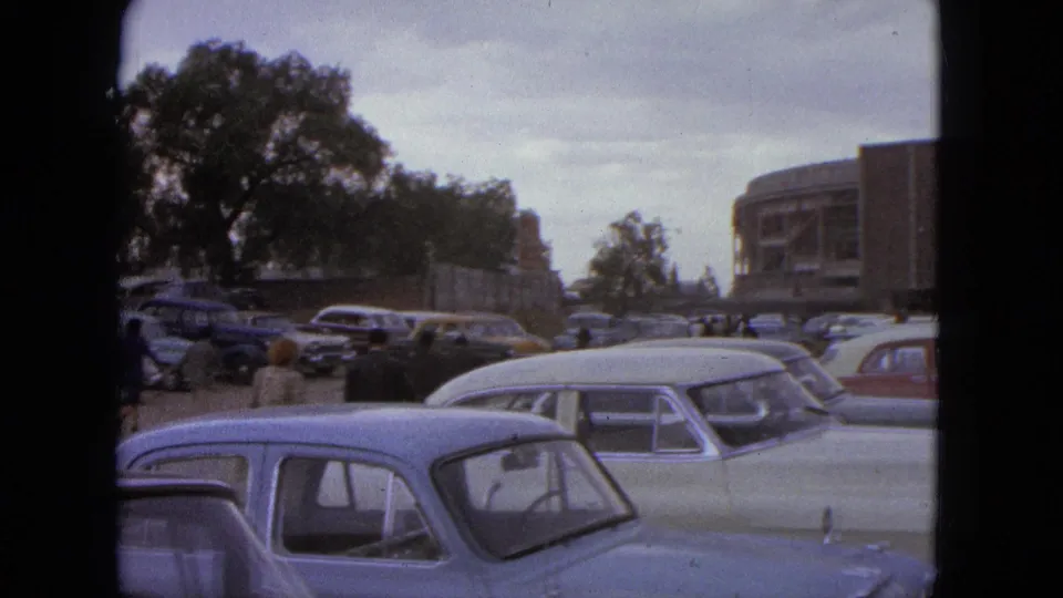 1962 Acapulco frame showing a wider parking area with classic cars, trees, people, and the edge of the TOREO building.