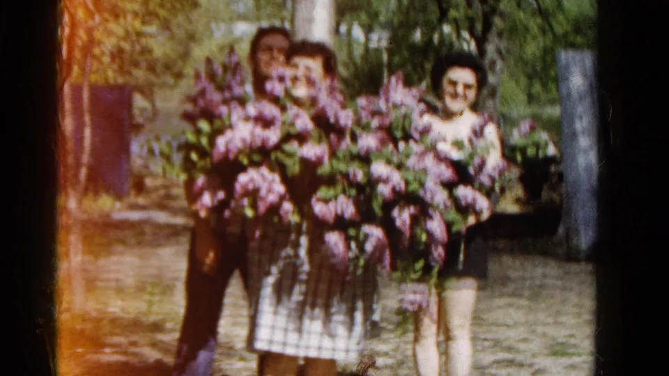 A 1961 Wauconda garden scene shows three adults smiling or posing behind oversized purple lilac bouquets.