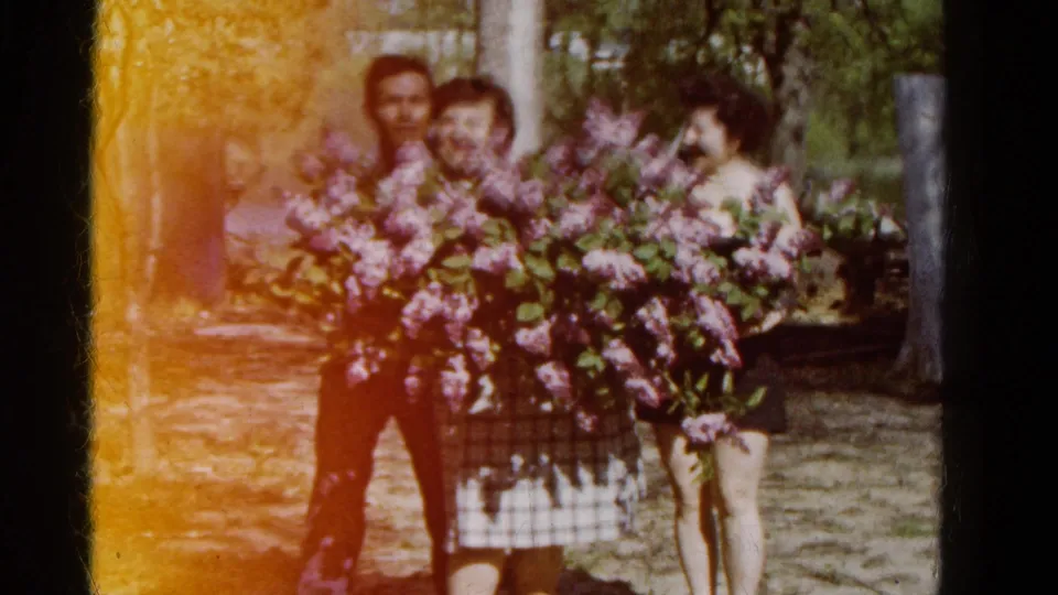 The home-movie frame shows three adults in a Wauconda garden with purple lilacs and visible 8mm light leak.