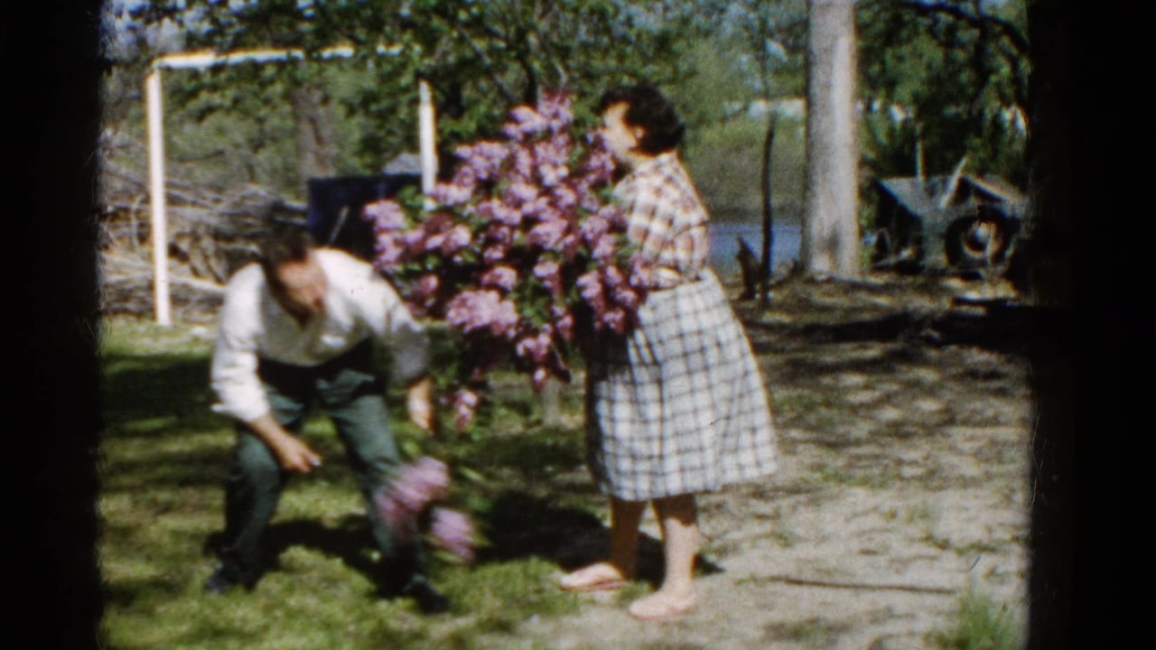 Man crouches beside a woman holding a large bouquet of purple flowers outdoors.