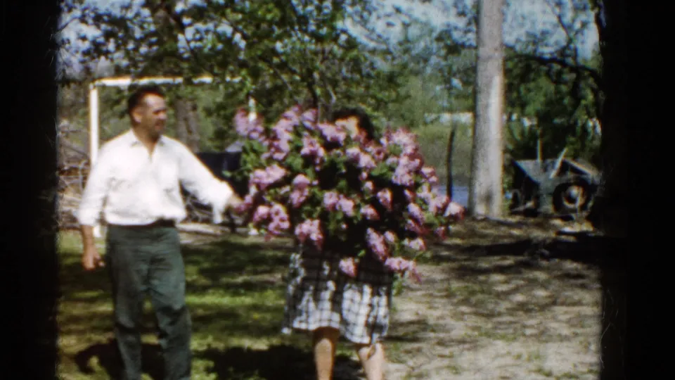 A man in a white shirt enters beside the woman with lilacs in the 1961 Wauconda backyard scene, with trees and old equipment behind them.
