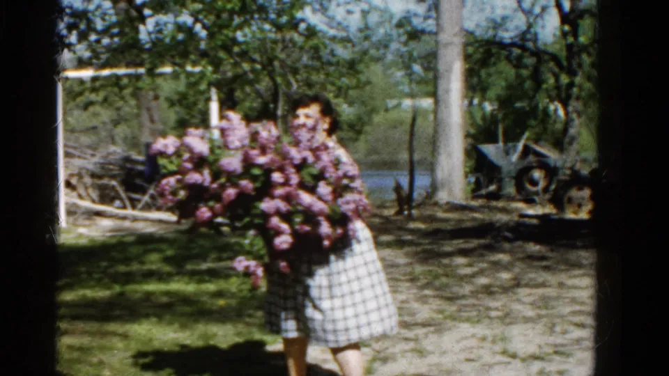 A woman in a patterned top and gingham skirt faces the camera while holding a large pink lilac bouquet in the Illinois yard.