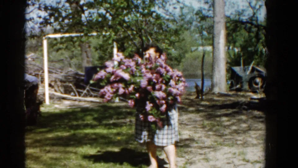 The bouquet partly obscures the woman as she stands in the Wauconda yard, with trees, water, and weathered equipment in the background.