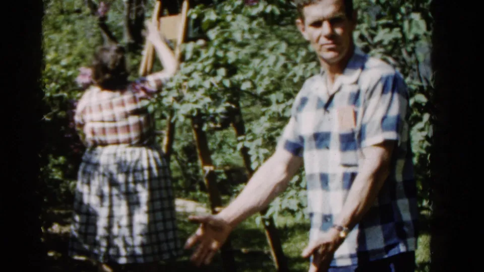A handheld 8mm frame shows the woman reaching into the flowering tree while the man gestures toward the camera in the yard.