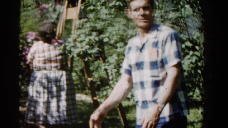 The man looks toward the camera in a softly focused 1961 home-movie frame as the woman remains near flowering branches behind him.