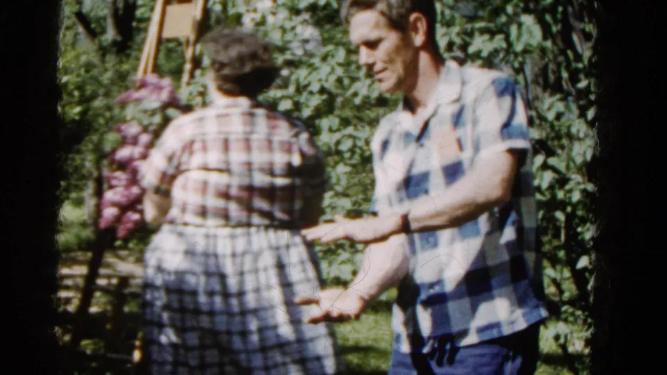In the Wauconda garden scene, the man extends his arm while the woman holding purple flowers moves toward foliage and a wooden ladder.