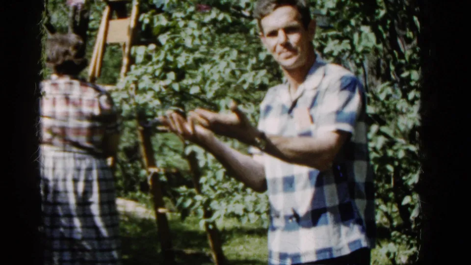 1961 home-movie frame showing the woman walking deeper into the garden as the man faces the camera and gestures with both hands.