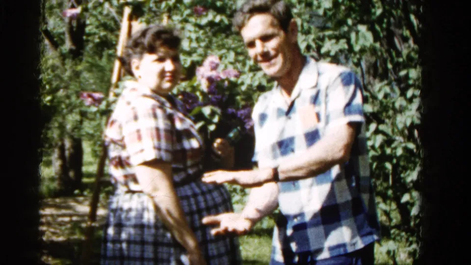 A woman in plaid and a man in a blue checkered shirt stand near lilacs in a 1961 Wauconda garden scene.