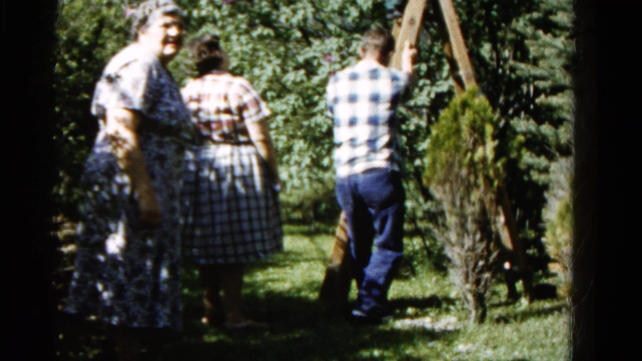 In 1961 Wauconda, Illinois home-movie footage, two women in patterned dresses watch a man near a wooden ladder in a green backyard.