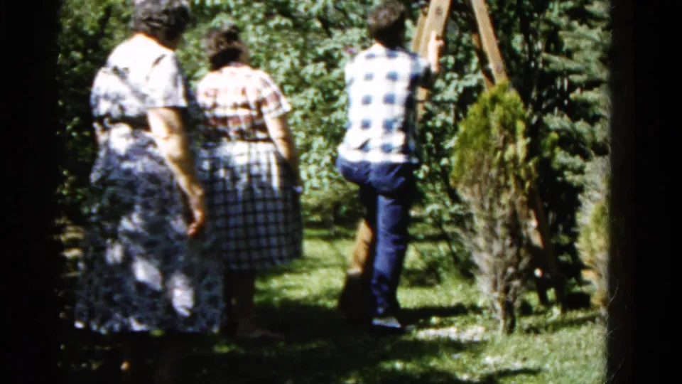 The man in a blue-and-white plaid shirt steps onto the wooden ladder as two women stand in the shaded 1961 Wauconda backyard.