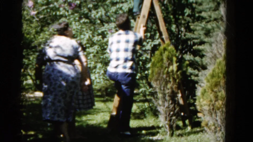 The man stands with one foot on the wooden ladder while two women remain nearby in the sun-dappled 1961 Wauconda yard.