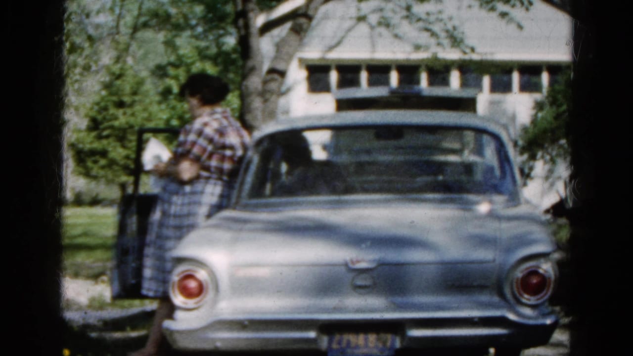 In 1961 Wauconda, Illinois, a woman in plaid clothing stands beside an open car door near a silver sedan and white garage.