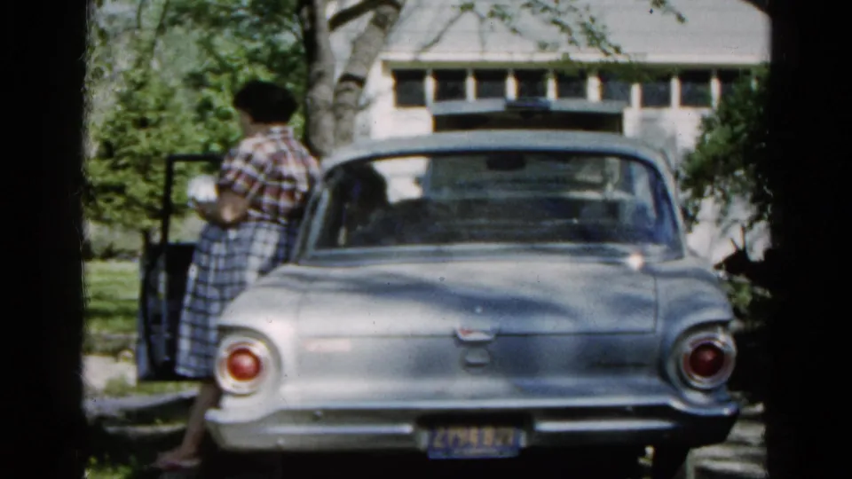 A 1961 Wauconda home-movie frame shows a woman near an open sedan door, with the car parked before a white garage.