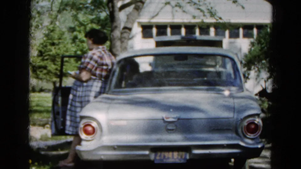 The woman remains beside the open car door in this 1961 Wauconda driveway view, with the sedan filling the foreground.