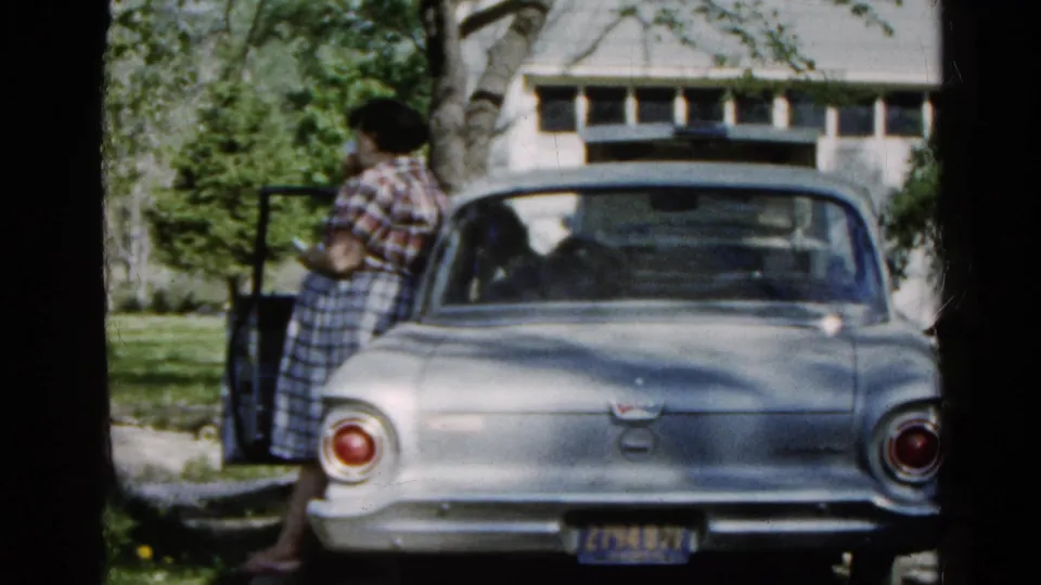 A silver early 1960s sedan sits in a Wauconda driveway while a woman stands near the open door, partly shaded by trees.