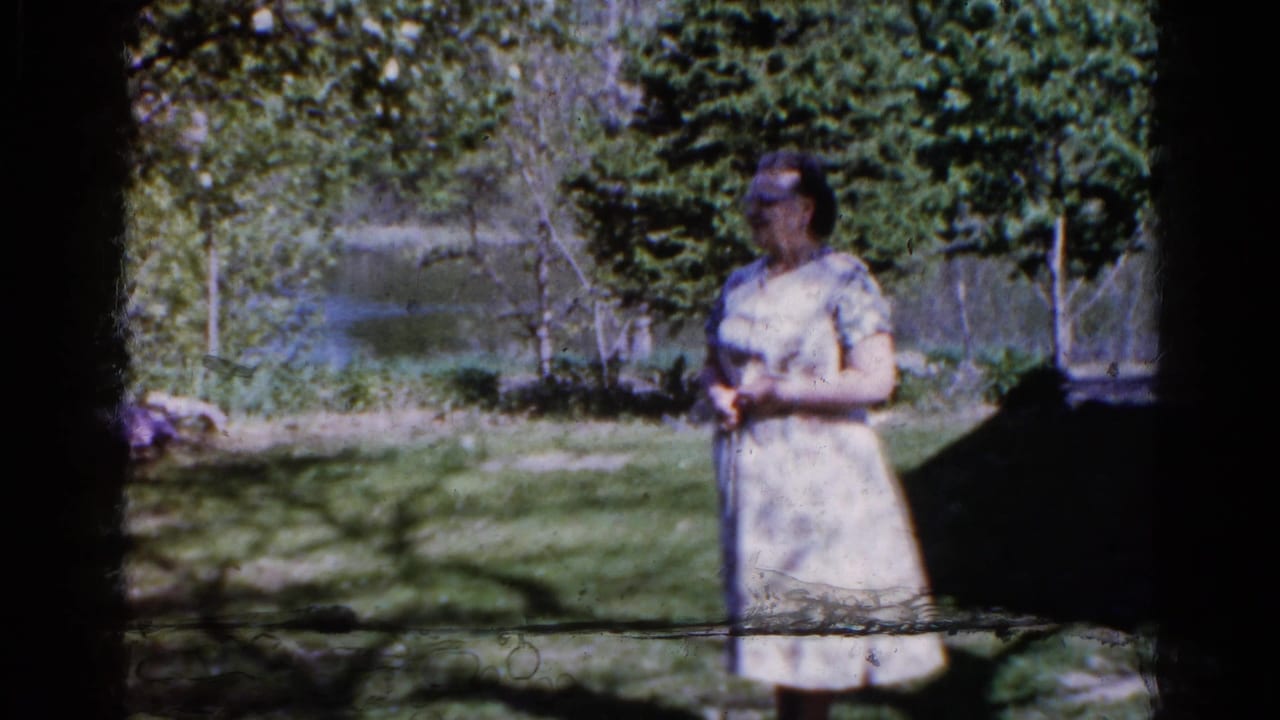 Woman in a light dress stands in a shaded yard near trees and water.