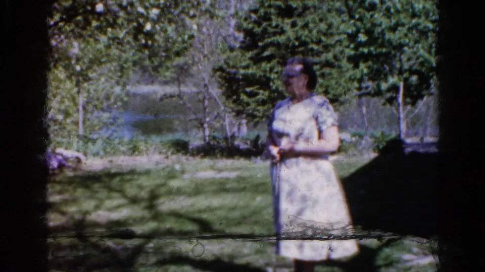 A woman stands outdoors in a Wauconda, Illinois yard in 1961, with trees, grass, and water visible behind her.