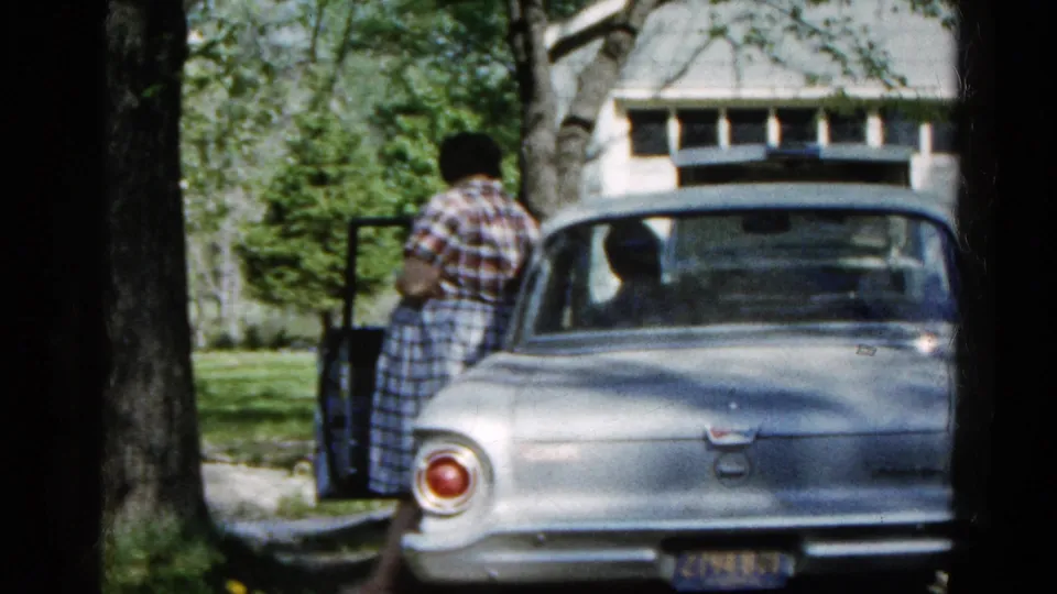 In 1961 Wauconda, a woman in plaid stands by the open driver's door of a silver sedan while a man sits inside.