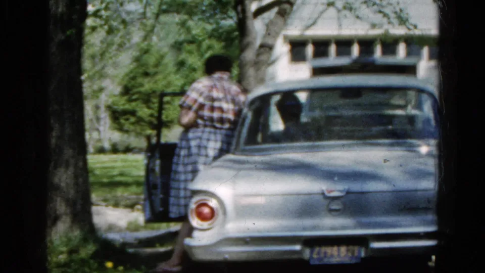 A woman stands beside the open driver's door of a silver mid-century sedan in a leafy Wauconda yard, with a house behind it.
