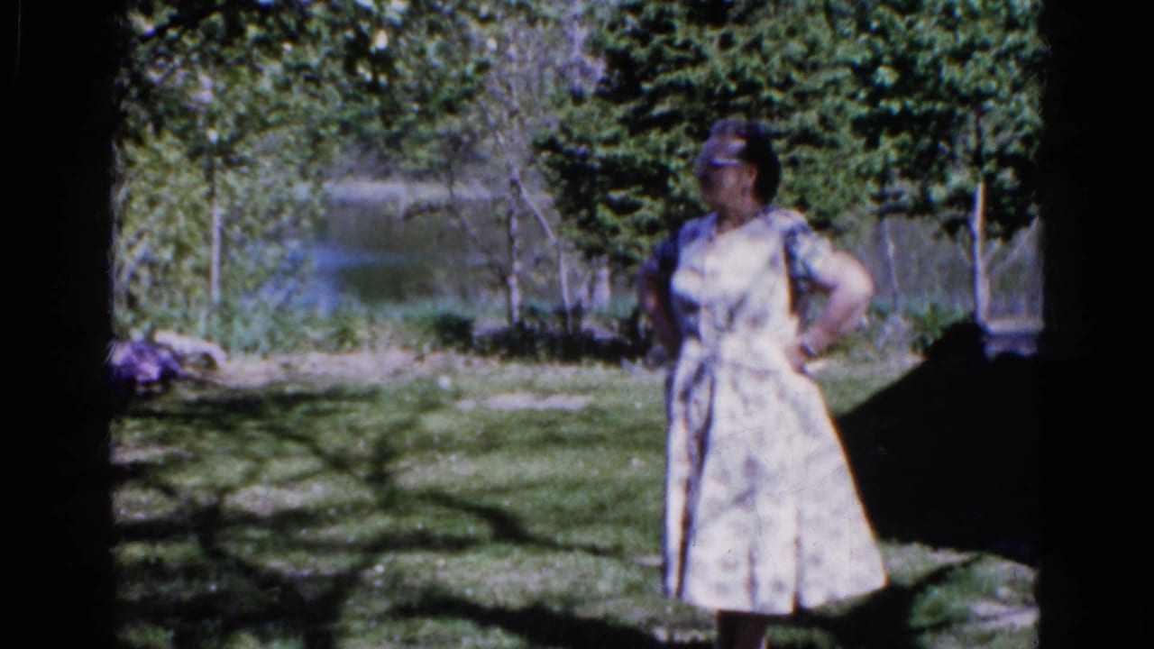 Woman in a light floral dress stands on grass beneath tree shade with water and trees behind her.