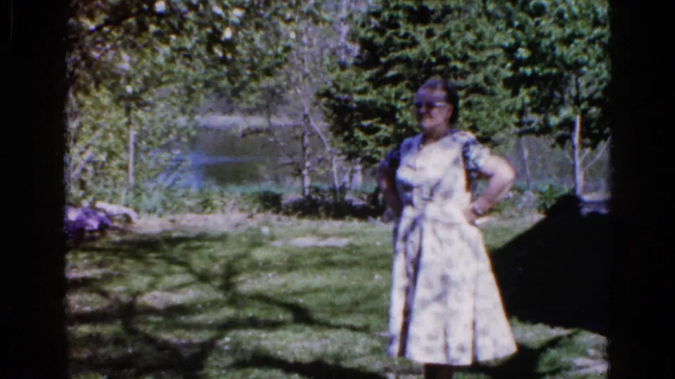 The woman remains posed under tree shade in 1961 Wauconda home-movie footage, with grass, water, and trees behind her.