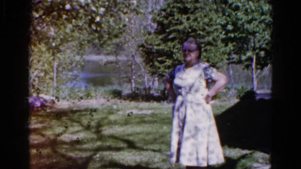 A woman in a pale patterned dress stands in a shaded grassy area near water in metadata-supported 1961 Wauconda, Illinois footage.