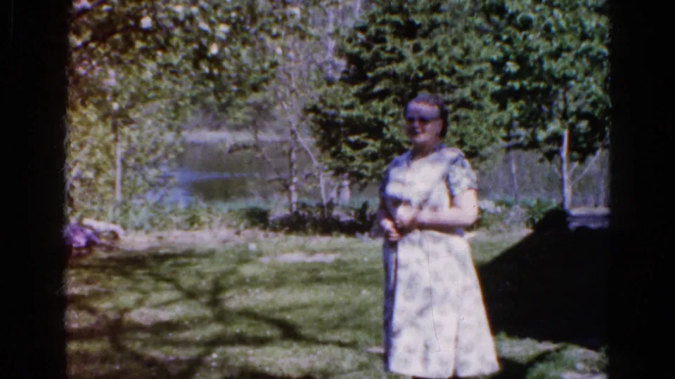 Near the end of the 1961 Wauconda clip, the woman shifts her hands while standing in tree shade beside a grassy lakeside yard.