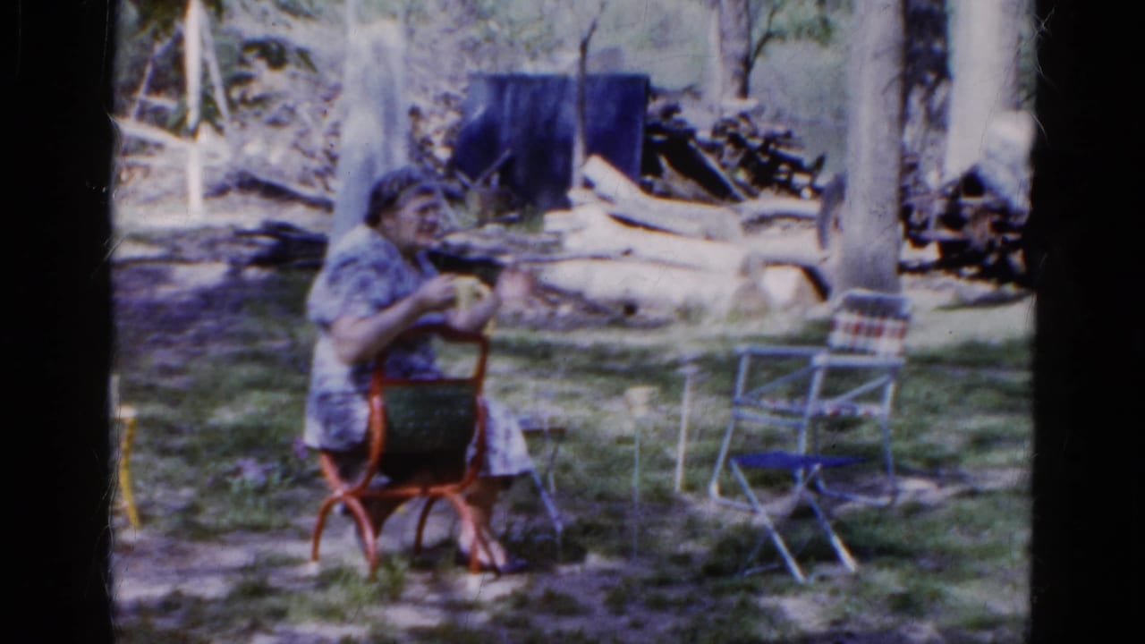 In Wauconda, Illinois, 1961, an older woman sits in a red metal chair in a shaded garden, with folding chairs and logs behind her.