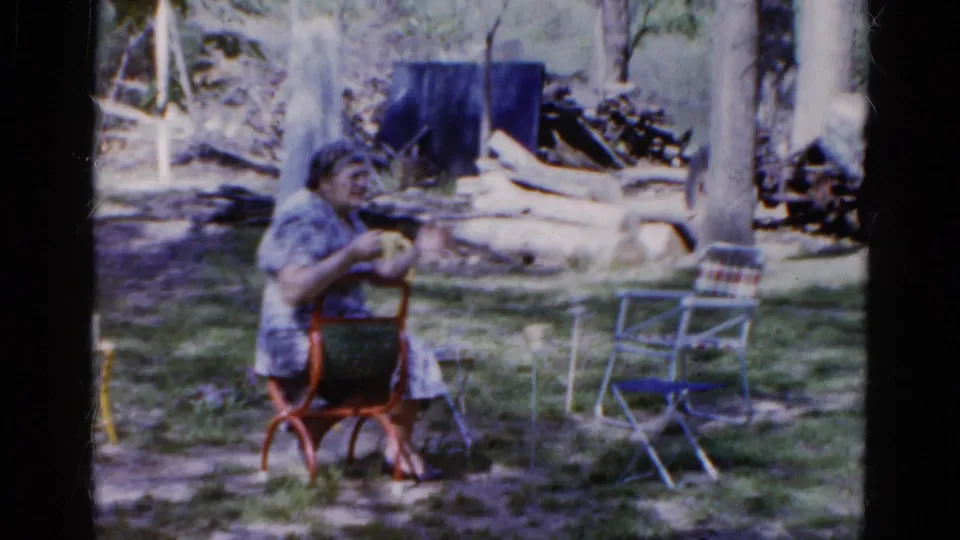 In Wauconda, Illinois, 1961, an older woman sits in a red metal chair in a shaded garden, with folding chairs and logs behind her.