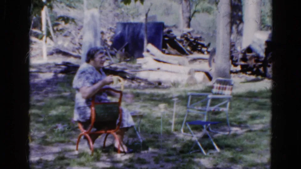 The seated woman appears to talk or gesture from a red metal chair in the 1961 Wauconda garden scene, with trees and chairs in the background.