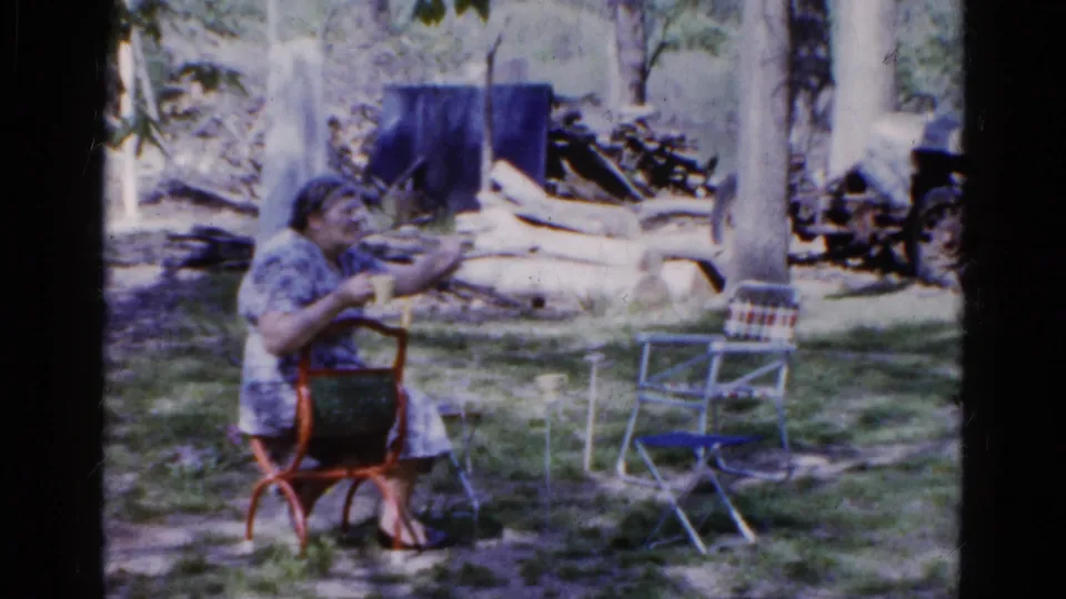 In the Wauconda home-movie garden scene, the older woman raises a hand as if speaking, seated near lawn chairs, trees, and stacked wood.