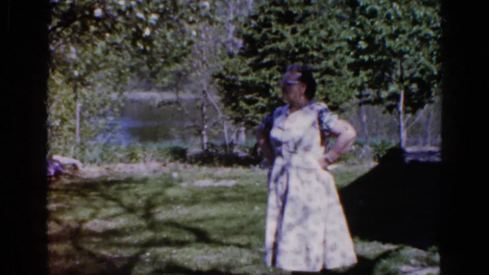 A second adult woman in a light patterned dress stands in the Wauconda yard, with trees, grass, and garden foliage visible behind her.