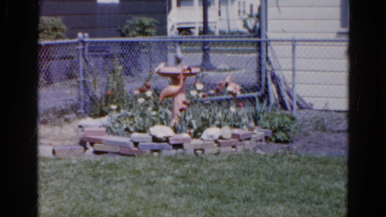 1961 Wauconda backyard view with a terracotta bird bath or fountain set inside a low brick flower bed near a chain-link fence.