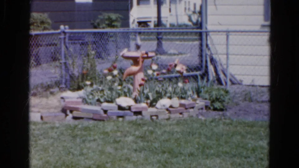 1961 Wauconda backyard view with a terracotta bird bath or fountain set inside a low brick flower bed near a chain-link fence.