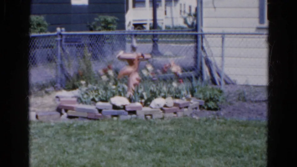A fixed 8mm view of a Wauconda backyard garden, with a terracotta bird bath, flowers, grass, and fencing behind it.