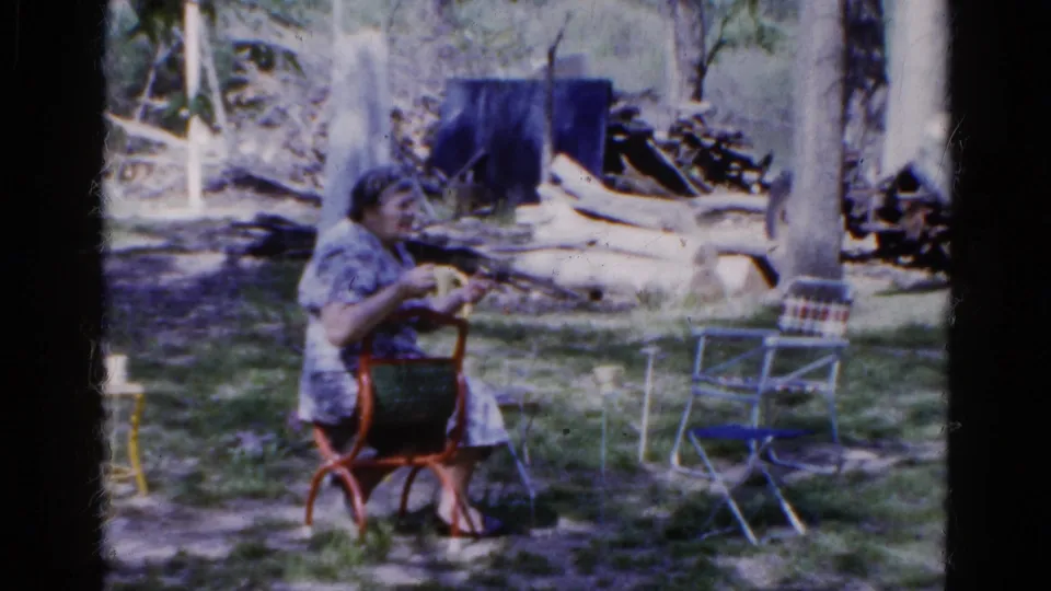 In a wooded outdoor area, a woman in a patterned dress sits in a red metal chair, apparently knitting or handling small objects.