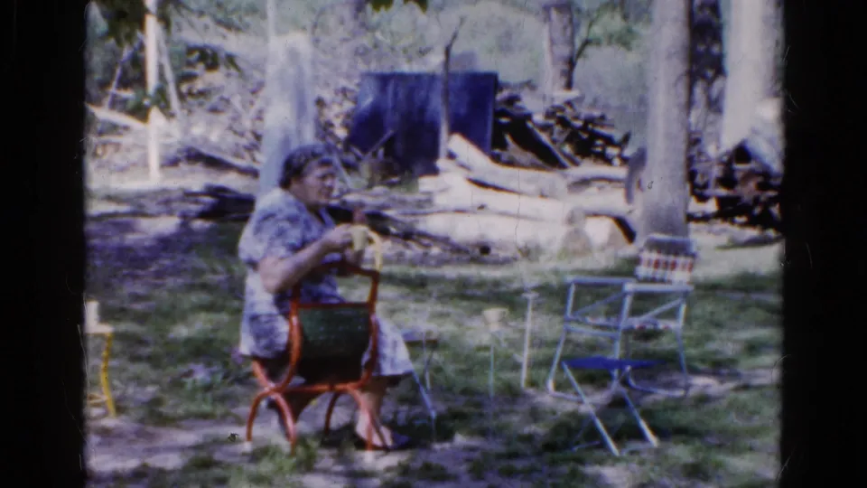 A woman sits outdoors in a red chair in 1961 Wauconda footage, with trees, grass, and nearby lawn chairs visible around her.
