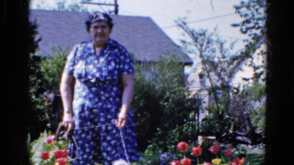 The woman faces the camera in the residential garden, with red tulips, dense greenery, and a light-colored house behind her.