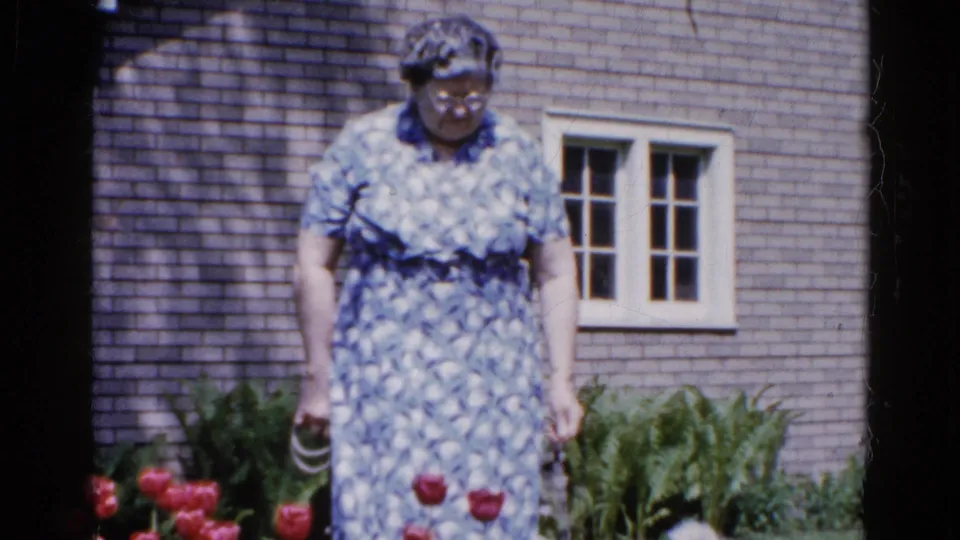 A woman stands near red tulips and broad garden leaves in front of a house wall with a multi-pane window.