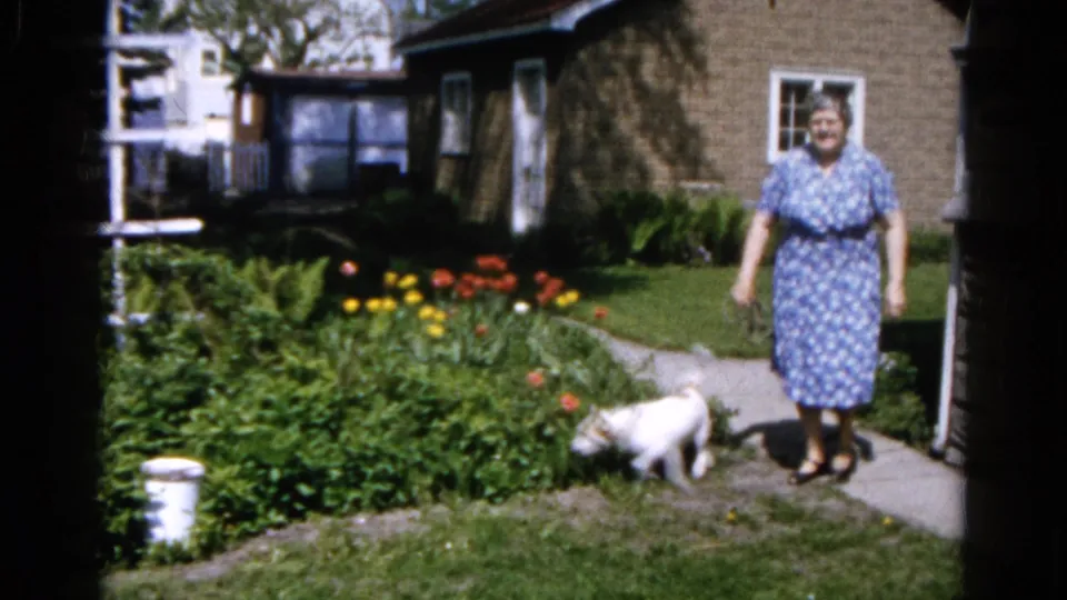The woman stands facing the camera in a Wauconda suburban yard as the small white dog sniffs near the flower bed and path.