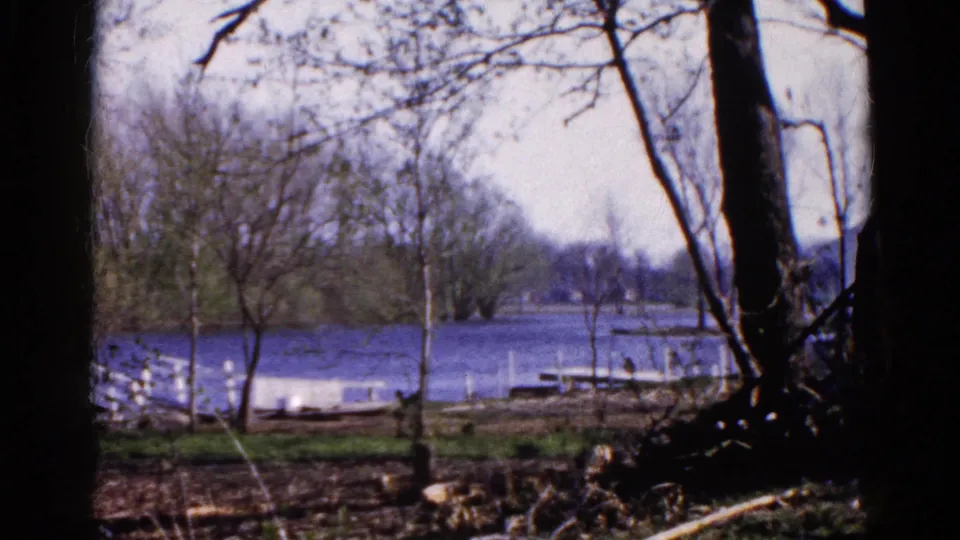 1961 8mm view in Wauconda, Illinois, looking through bare trees toward a calm blue lake and small docks.