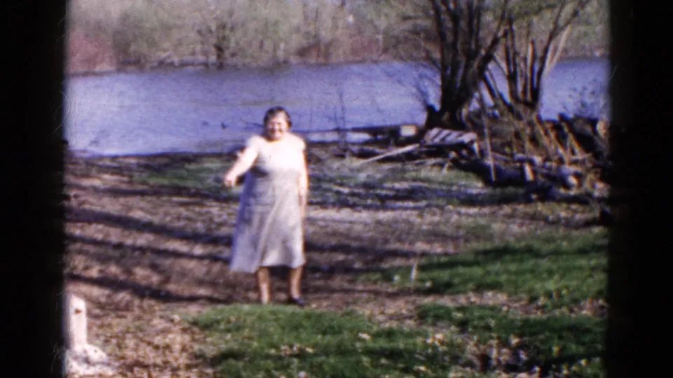 The adult woman faces the camera again near the Wauconda lake, framed by grass, dirt ground, logs, trees, and water.