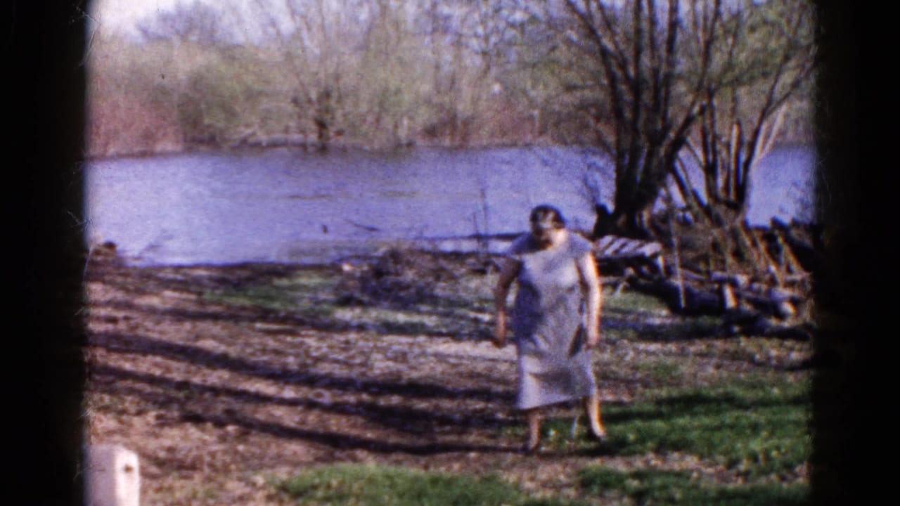 Woman in a pale dress standing on a grassy riverbank near bare trees and water.