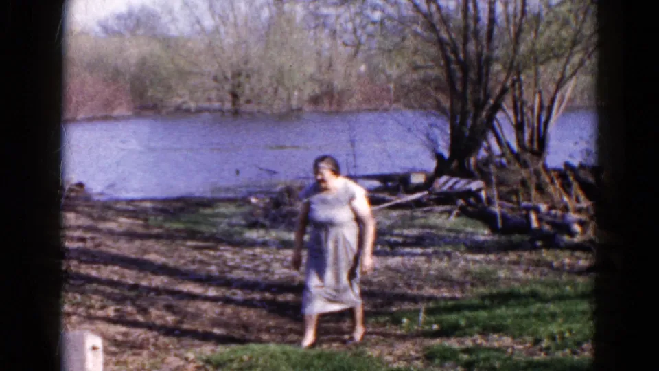 The woman faces the camera on a grassy riverbank in 1961 Wauconda footage, with water, leafless trees, and stacked logs visible behind her.