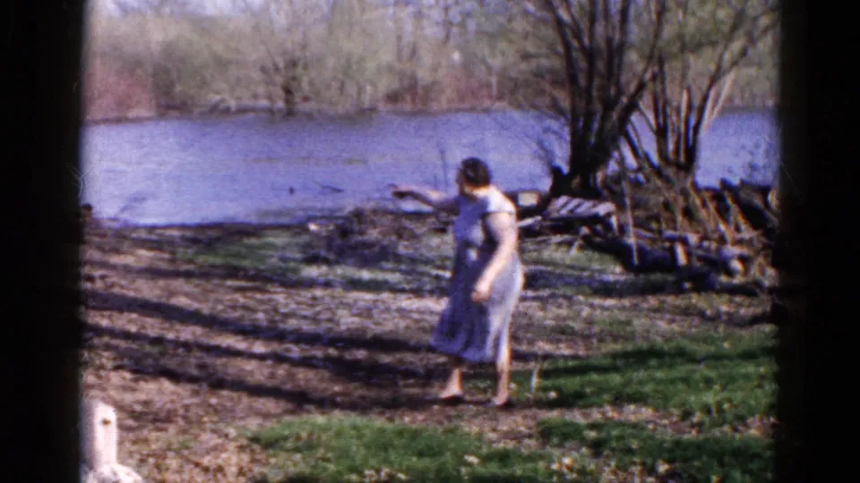 The woman walks along the riverbank in the 1961 Wauconda home movie, seen in profile against water, bare trees, grass, and muddy ground.
