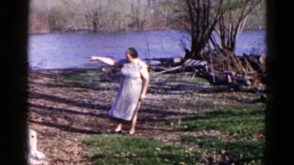 In the final frame, the woman points toward the river from the grassy bank, with Wauconda water, trees, logs, and soft 8mm color visible.