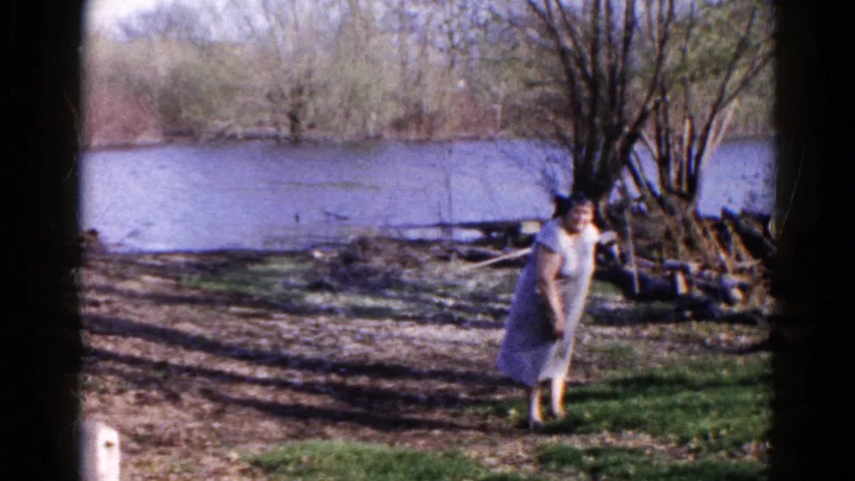 A woman in a light dress bends slightly beside a riverbank in the supplied 1961 Wauconda home-movie scene.