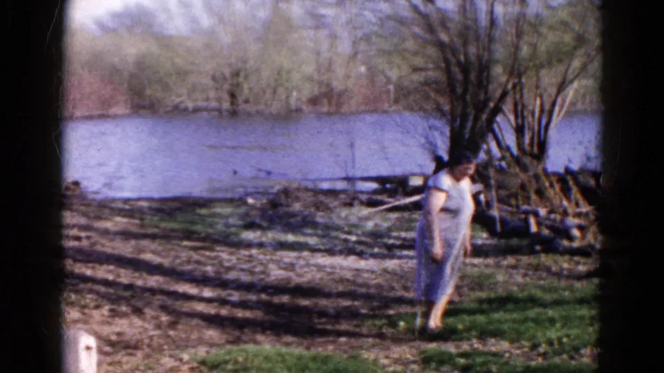 The woman walks toward the camera along uneven riverside ground in 1961 Wauconda, Illinois footage.
