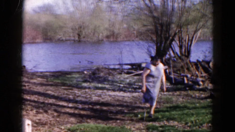 A woman in a pale dress steps along the grassy riverbank in the Wauconda, Illinois home-movie clip.